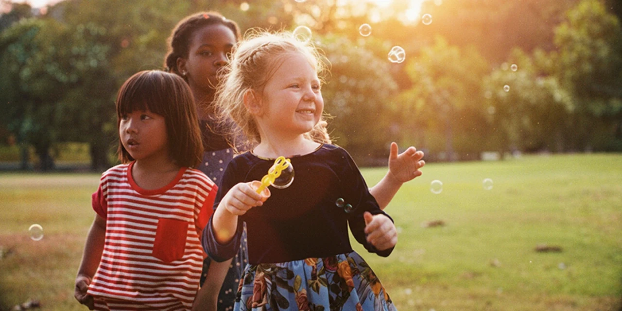 Drei Kinder spielen an einem sonnigen Nachmittag auf einer Wiese mit Seifenblasen. Ein blondes M&auml;dchen h&auml;lt einen Seifenblasenstab in der Hand und l&auml;chelt, w&auml;hrend zwei weitere Kinder hinter ihr stehen.